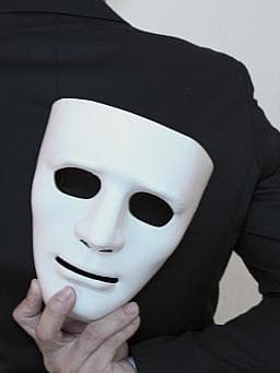 Eine Person in einem schwarzen Anzug hält eine weiße Maske hinter ihrem Rücken. Die Maske ist teilweise verdeckt und die Person ist von hinten zu sehen.