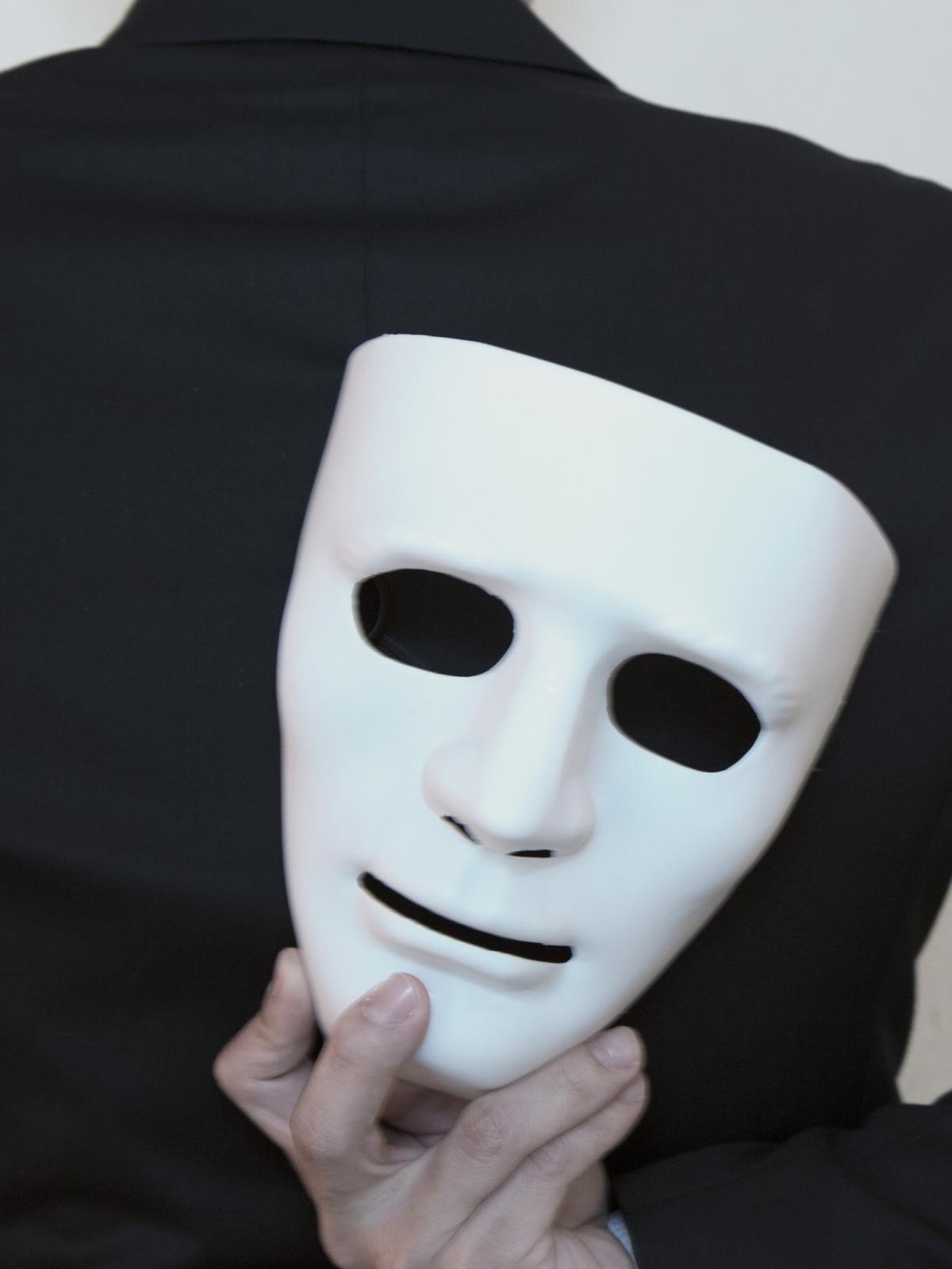Eine Person in einem schwarzen Anzug hält eine weiße Maske hinter ihrem Rücken. Die Maske ist teilweise verdeckt und die Person ist von hinten zu sehen.