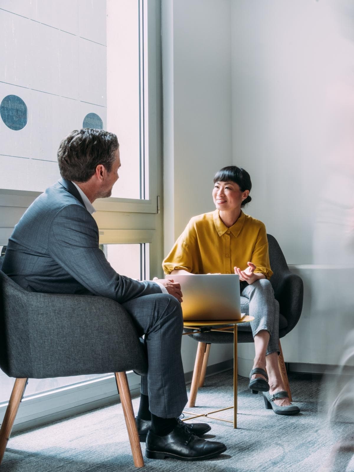 Zwei Personen sitzen und unterhalten sich in einem Büroflur, die Frau mit einem Laptop. Unscharfe Figuren suggerieren Bewegung und vermitteln eine geschäftige, dynamische Atmosphäre.