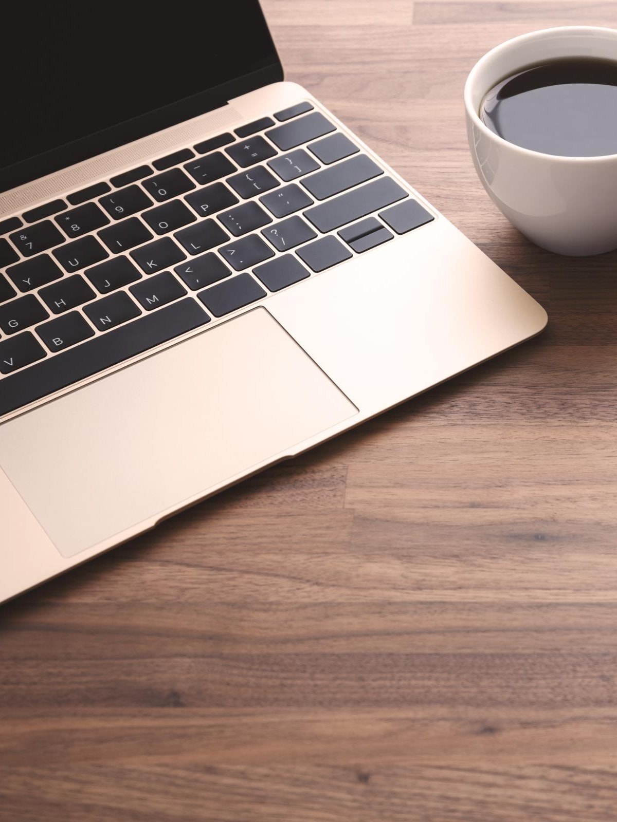 Auf einem hellbraunen Holztisch stehen ein Laptop, eine Tasse Kaffee, ein Stift und ein Notizbuch.