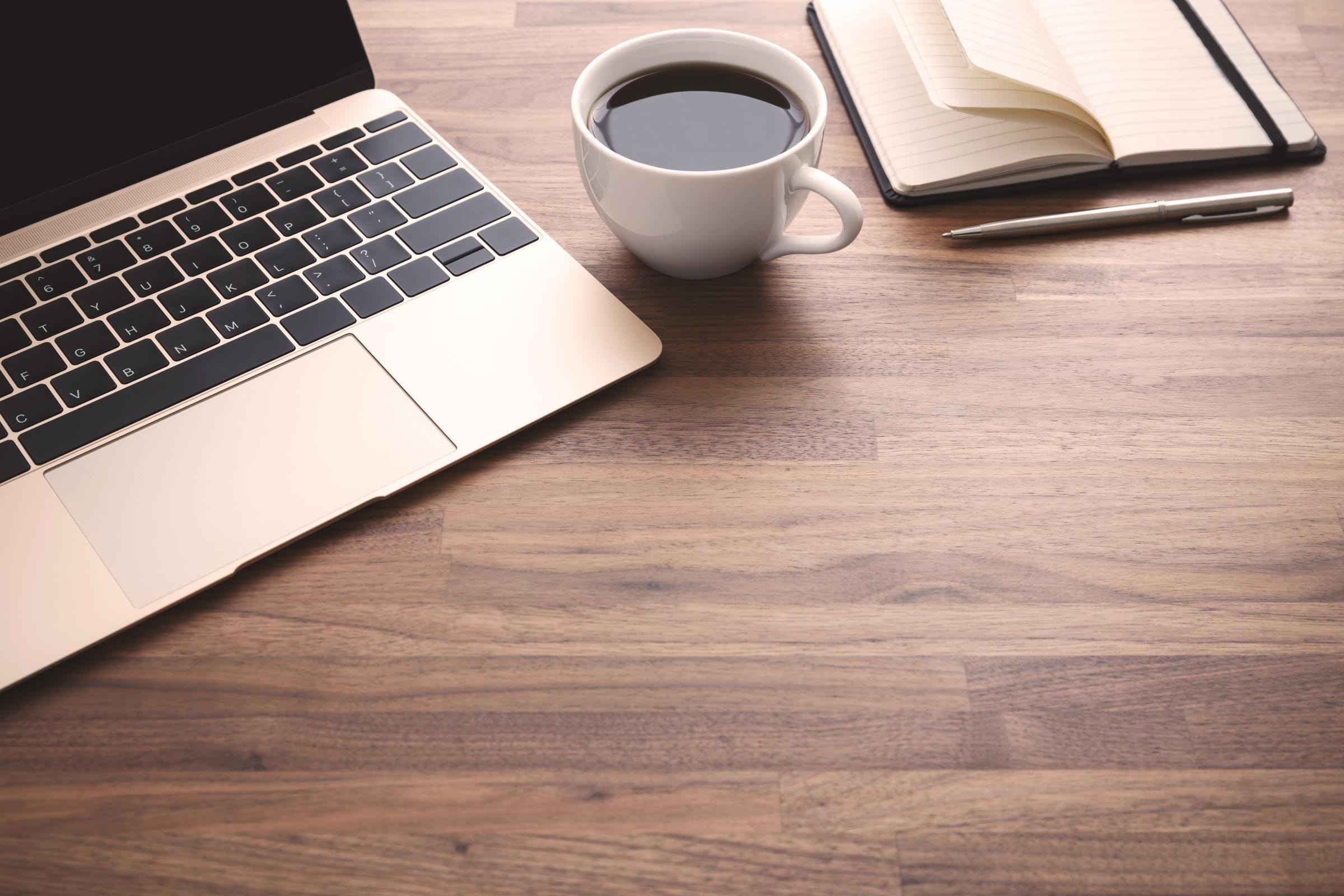 Auf einem hellbraunen Holztisch stehen ein Laptop, eine Tasse Kaffee, ein Stift und ein Notizbuch.