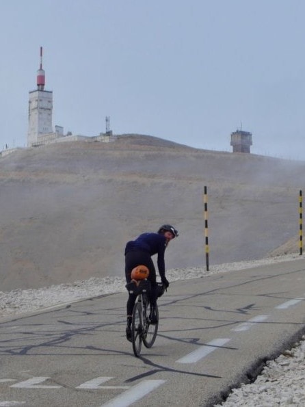 Man sieht den Extremsportler Jonas Deichmann auf einem Rennrad einen Berg in einer sehr kargen Landschaft auf einer Straße hinauffahren. Er dreht sich beim Fahren um und schaut in die Kamera. Oben am Berg steht eine Art Leuchtturm.