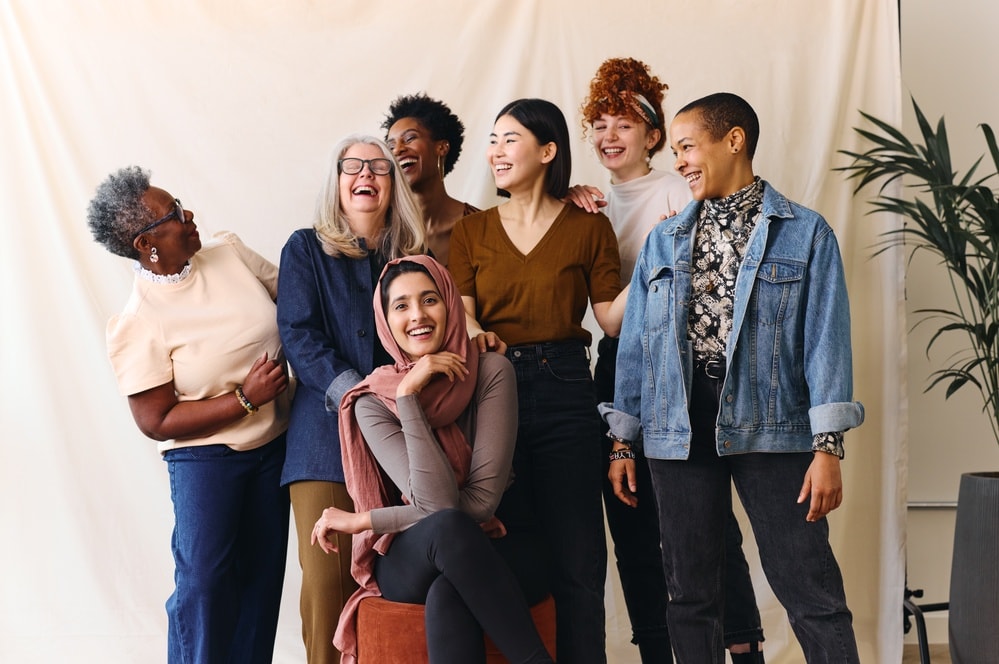 Sieben Frauen unterschiedlicher Herkunft stehen zusammen, lächeln und lachen. Sie strahlen in einer ungezwungenen, freundlichen Umgebung ein Gefühl der Zusammengehörigkeit und Freude aus. Sie repräsentieren gründende Frauen.