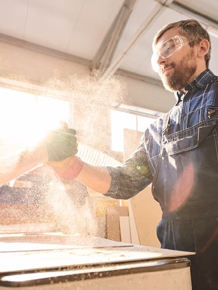 Zwei männliche Handwerker in Schutzkleidung mit Schutzbrillen stehen sich in einer Werkstatt gegenüber und geben sich einen Handschlag. Die Sägespähne fliegen durch die Luft und die Sonne scheint durch das Fenster.