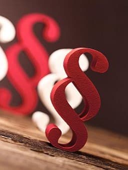 Mehrere rot-weiße Paragrafzeichen in 3D stehen gemischt auf einer Holzfläche.