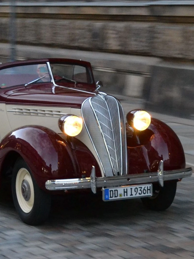 Ein klassisches rot-weißes Cabriolet aus den 1930er Jahren fährt auf einer gepflasterten Straße entlang einer historischen Steinmauer. Das Auto hat runde Scheinwerfer und ein markantes Chromgitter an der Front.