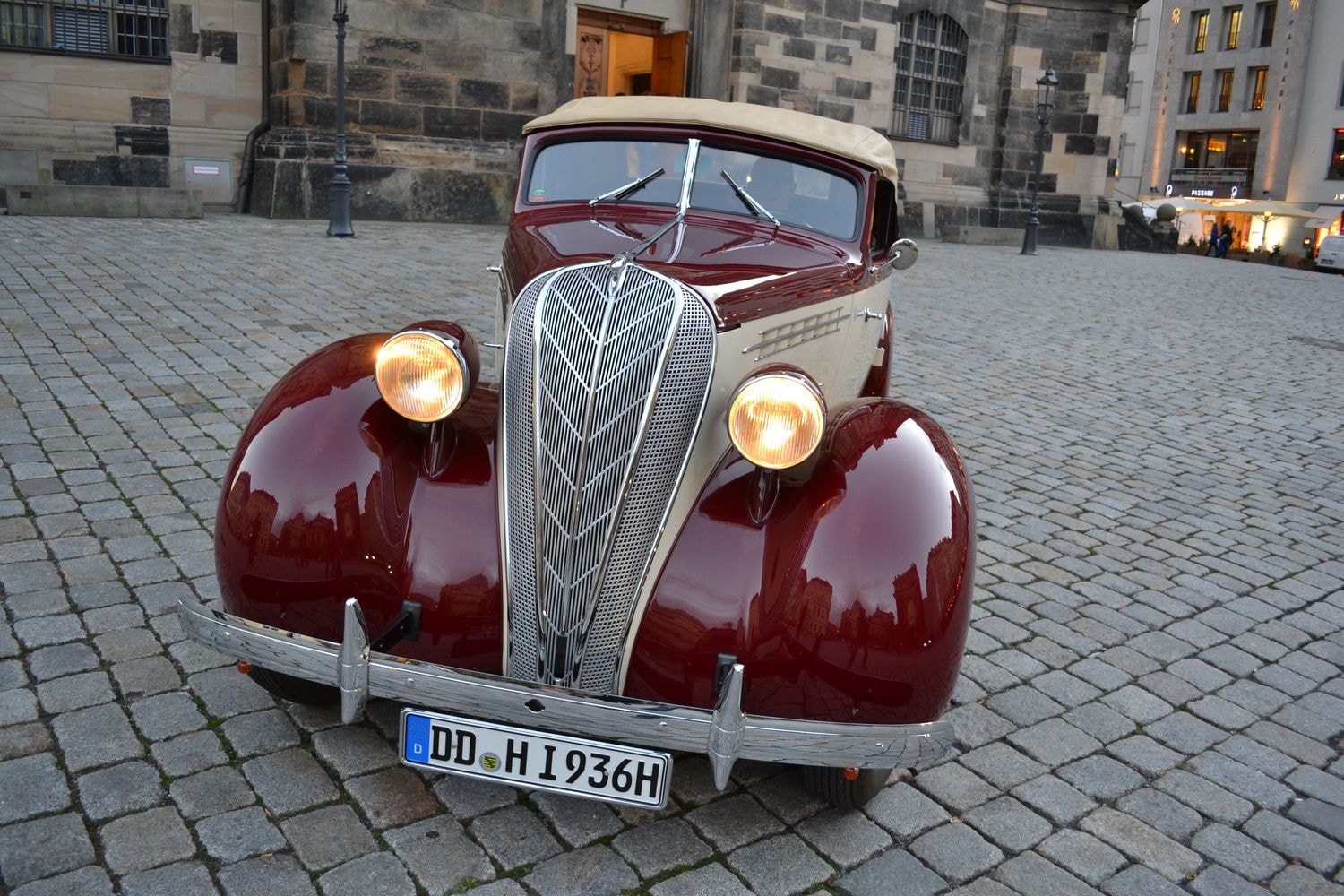 Ein Oldtimer ist auf einer Kopfsteinpflasterstraße vor einem alten Gebäude geparkt. Das Auto hat ein markantes, klassisches Design mit einem auffälligen Kühlergrill und runden Scheinwerfern.