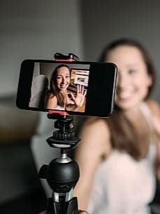 Lächelnde Influencerin sitzt vor ihrem PC und hält eine Vlogging-Kamera in der Hand und filmt sich selber