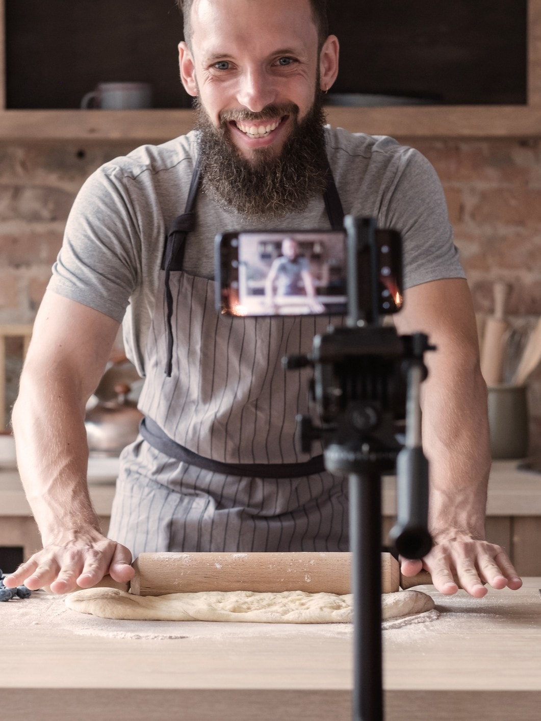 Ein Mann mit Bart rollt Teig in einer Küche aus, während ein Smartphone auf einem Stativ den Vorgang filmt. Im Hintergrund sind eine Backsteinwand, Küchenschränke und Küchenutensilien zu sehen.
