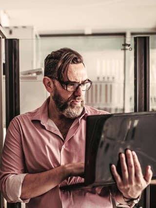 Mann mit Brille und Bart arbeitet an einem Laptop in einem Serverraum. Im Hintergrund sind mehrere Server-Racks zu sehen. Das Bild spiegelt sich in der Glaswand neben ihm.