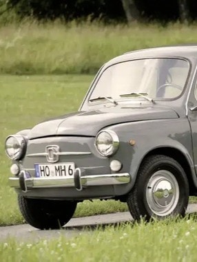 Ein grauer, klassischer Fiat 600, geparkt auf einer Wiese mit Bäumen im Hintergrund. Das Auto hat runde Scheinwerfer, Chromstoßstangen und kleine Räder.