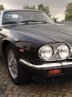 Ein klassischer schwarzer Jaguar XJ-S, geparkt auf einer Kopfsteinpflasterfläche. Das Auto hat markante runde Scheinwerfer, ein schlankes und aerodynamisches Design sowie Chromakzente am Kühlergrill und Stoßfänger.