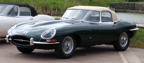 Ein klassisches, dunkelgrünes Jaguar E-Type Cabriolet mit cremefarbenem Verdeck und Speichenrädern, das im Freien neben einem anderen Oldtimer geparkt ist.