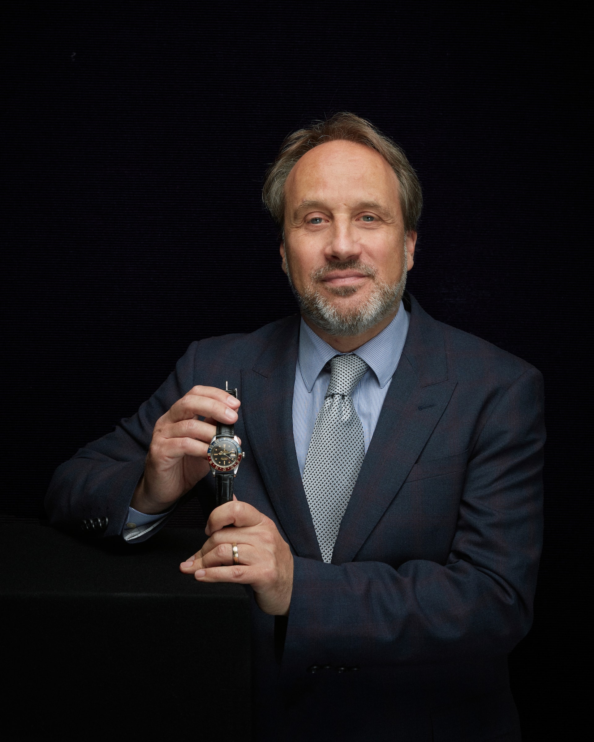 Jonathan Darracot, Global Head of Watches bei Bonhams, trägt einen dunkelblauen Anzug und hält eine Armbanduhr in die Kamera