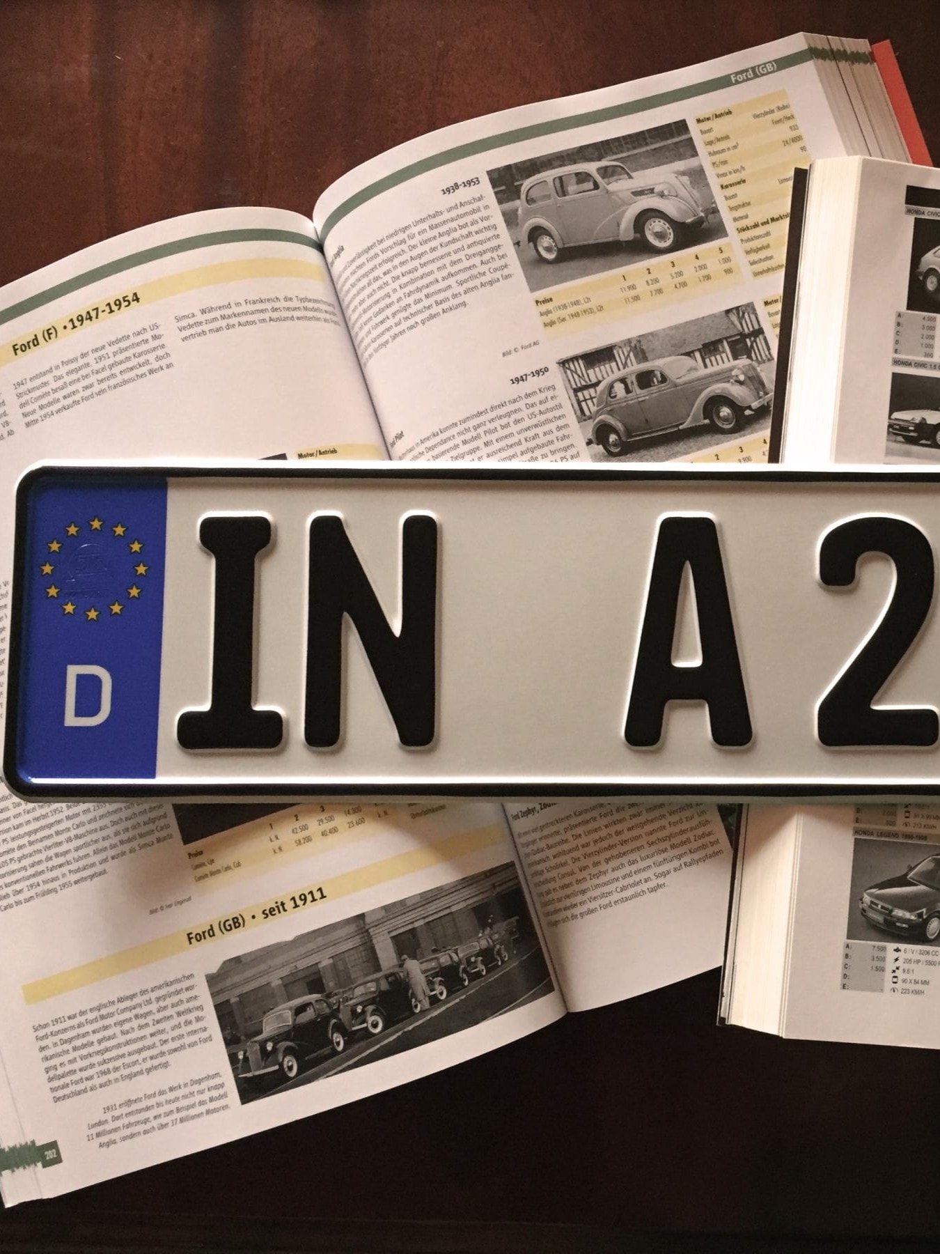 Ein deutsches Autokennzeichen mit der Aufschrift 'IN A 2563H' liegt auf einem aufgeschlagenen Automagazin, das verschiedene Autos und deren Spezifikationen zeigt.