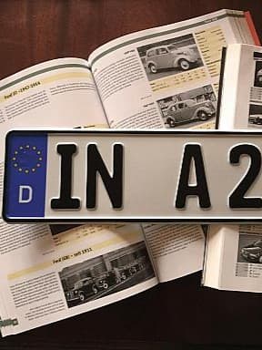 Ein deutsches Autokennzeichen mit der Aufschrift 'IN A 2563H' liegt auf einem aufgeschlagenen Automagazin, das verschiedene Autos und deren Spezifikationen zeigt.