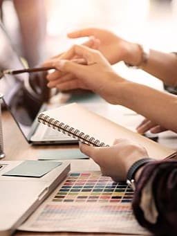 Zwei Personen arbeiten an einem Schreibtisch. Eine Person zeigt auf einen Laptop-Bildschirm.