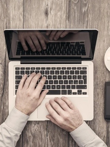 Die Hände einer Person tippen auf einem Laptop. Rechts auf dem Tisch sind eine Kaffeetasse, ein Tablet, ein Smartphone und ein Notizbuch mit einem Stift.