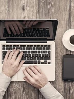 Die Hände einer Person tippen auf einem Laptop. Rechts auf dem Tisch sind eine Kaffeetasse, ein Tablet, ein Smartphone und ein Notizbuch mit einem Stift.