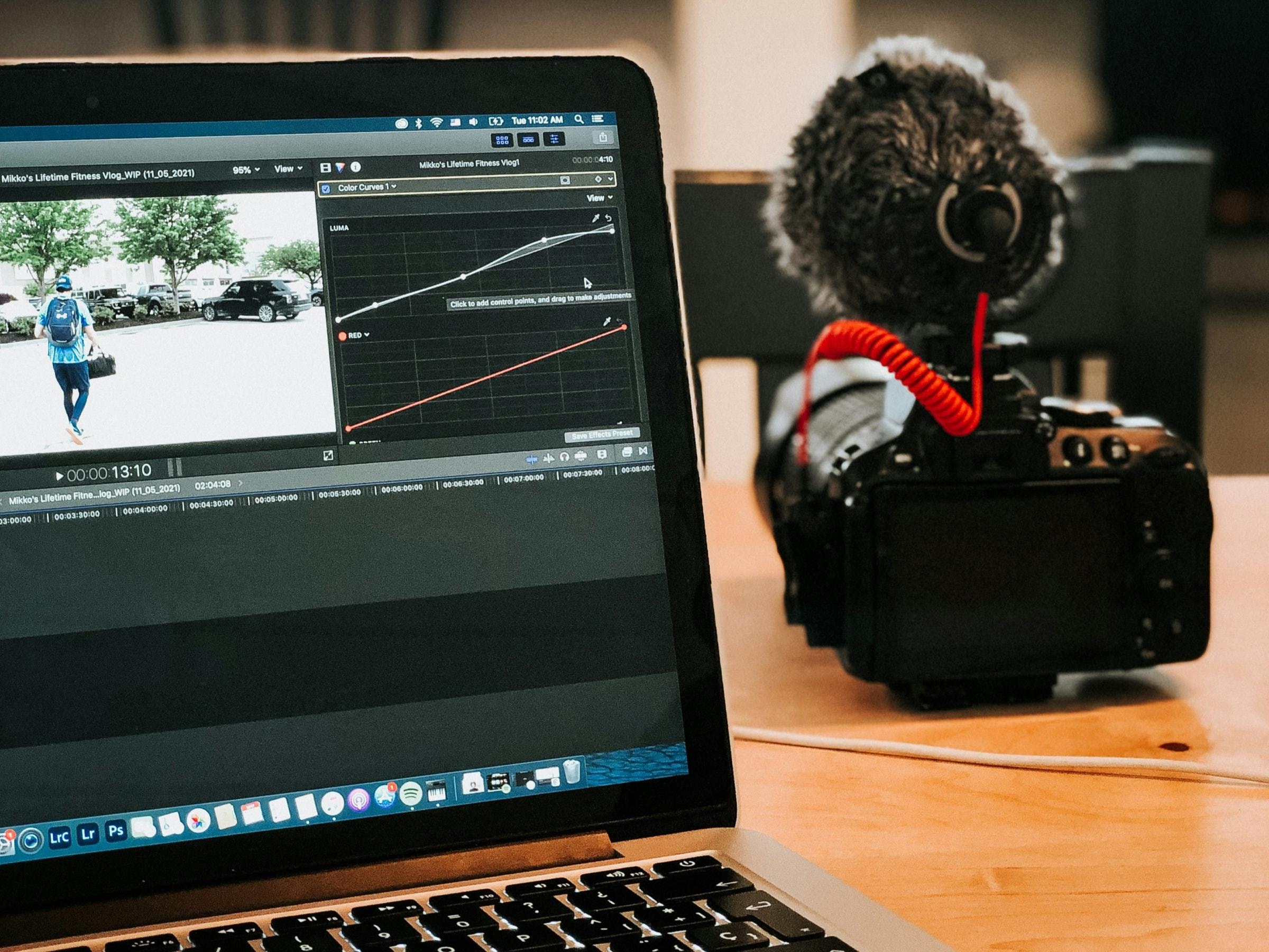 Auf einem Holztisch steht ein Laptop, auf dem eine Videobearbeitungssoftware läuft, daneben eine Kamera mit Mikrofon. Die Szene vermittelt den Eindruck eines kreativen Arbeitsbereichs.