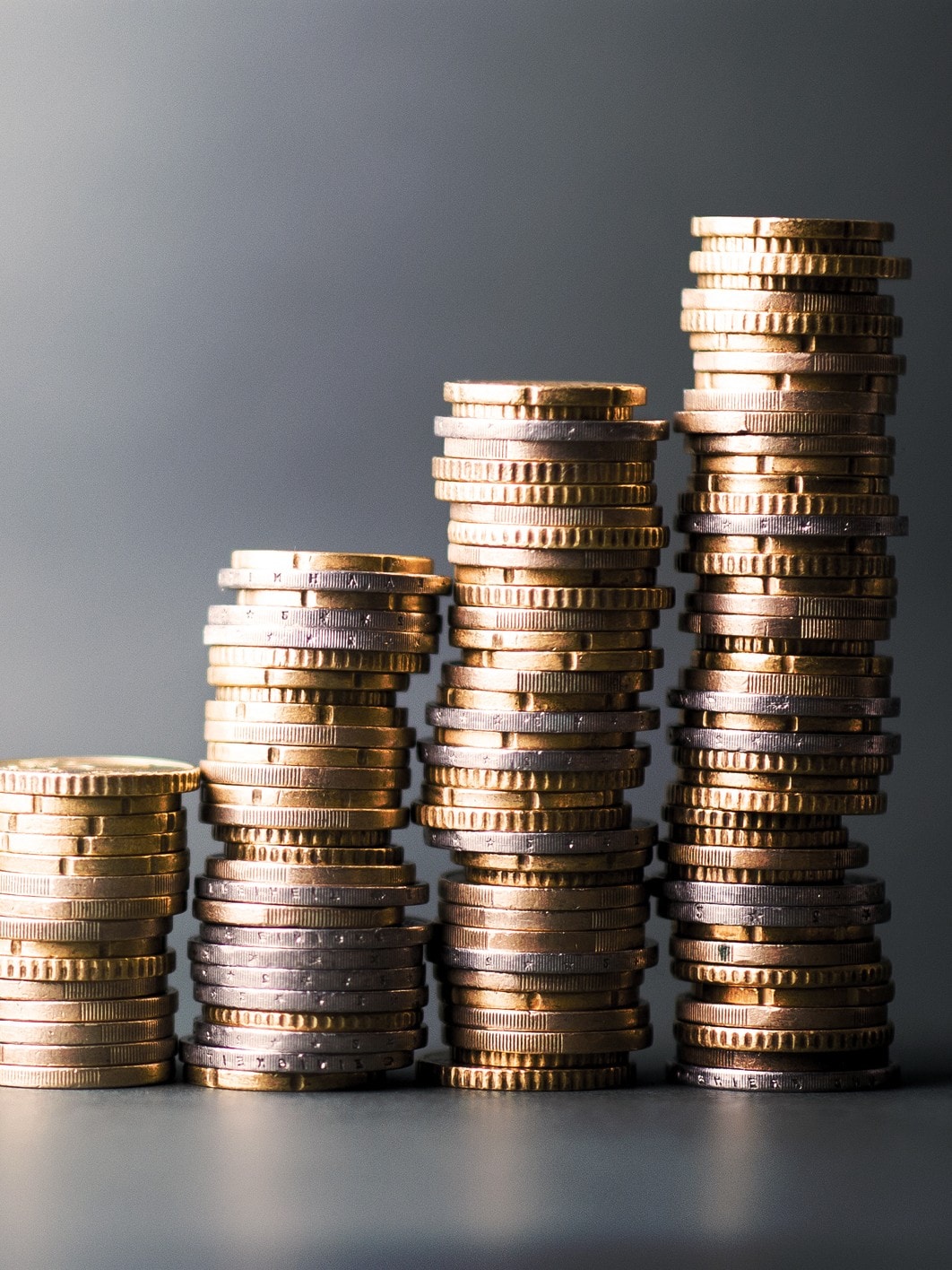 Gestapelte Münzen in verschiedenen Höhen, die von links nach rechts ansteigen. Die Münztürme symbolisieren ein wachsendes Vermögen. Hintergrund ist schlicht und grau gehalten.