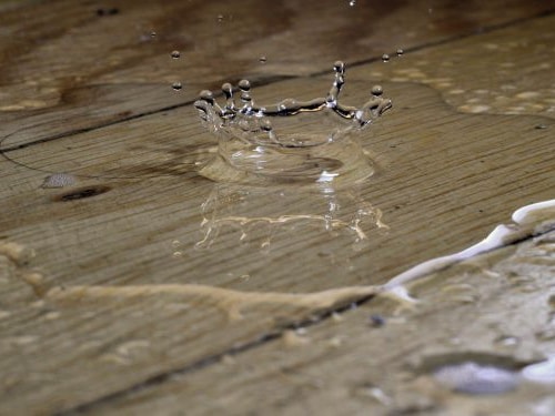 Nahansicht auf einen Terrassenboden aus Holz auf dem das Wasser steht. Ein großer Regentropfen fällt in die Mitte des Bildes