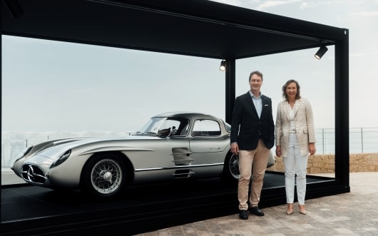 Ein eleganter, silberner Oldtimer, der im Freien in einer Glasvitrine ausgestellt ist. Ein Mann in einem dunklen Blazer und eine Frau in einem hellen Kostüm stehen lächelnd daneben und strahlen Stolz aus.