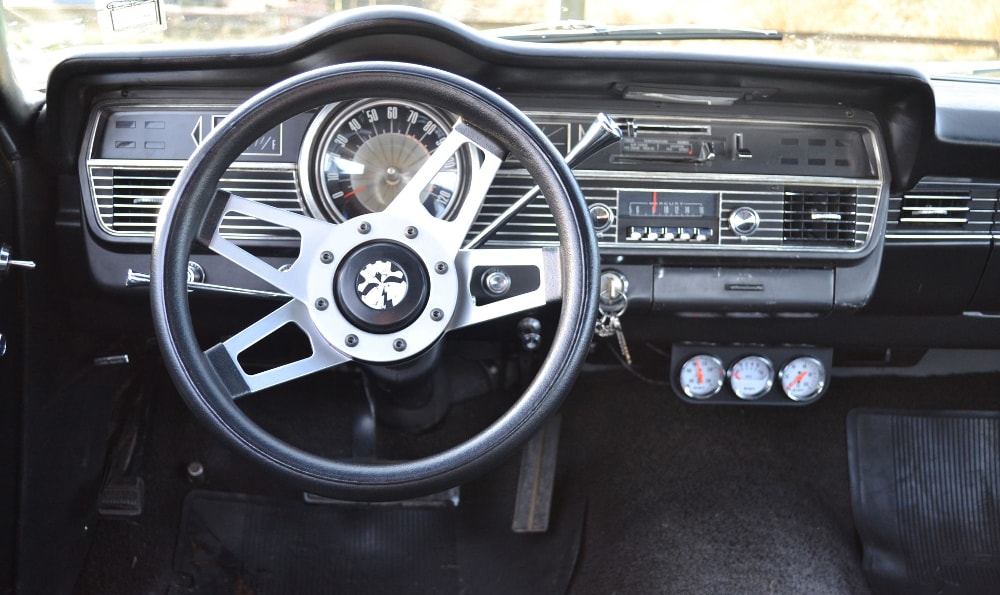 Das Bild zeigt das Armaturenbrett eines klassischen Autos mit einem Dreispeichen-Lenkrad und einem Tachometer bis 120 mph. Es gibt verschiedene Bedienelemente und Anzeigen, darunter eine analoge Uhr und ein Radio.