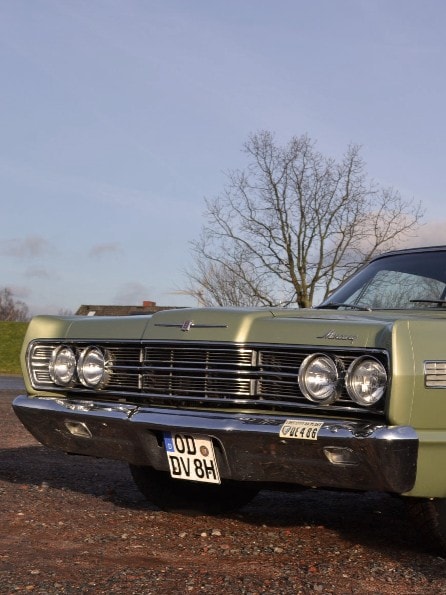 Ein klassischer grüner Ford Galaxie 500 ist auf einer Kiesfläche geparkt. Der Himmel ist klar und im Hintergrund sind einige Bäume zu sehen. Das Auto hat Chromdetails, runde Scheinwerfer und ein schwarzes Dach.