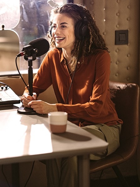 An einem Tisch sitzen ein Mann und eine Frau mit Kopfhörern, beide scheinen in ein Gespräch vertieft zu sein, das sie im Rahmen einer Podcastaufzeichnung über Micros aufnehmen.