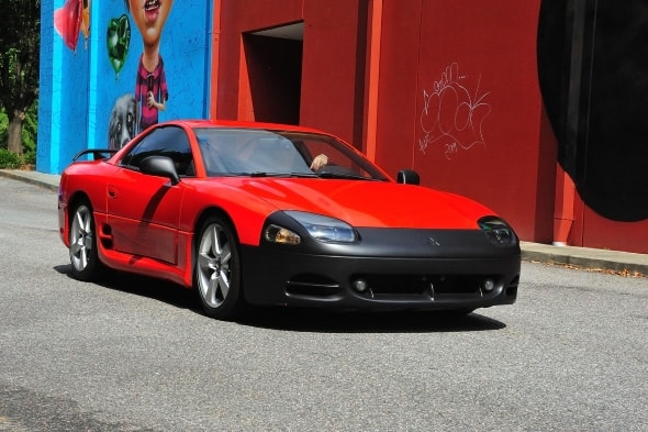 Ein rot-schwarzer Mitsubishi 3000GT Spyder fährt auf einer geteerten Straße. Man erkennt einen Fahrer hinter dem Steuer des Oldtimer.