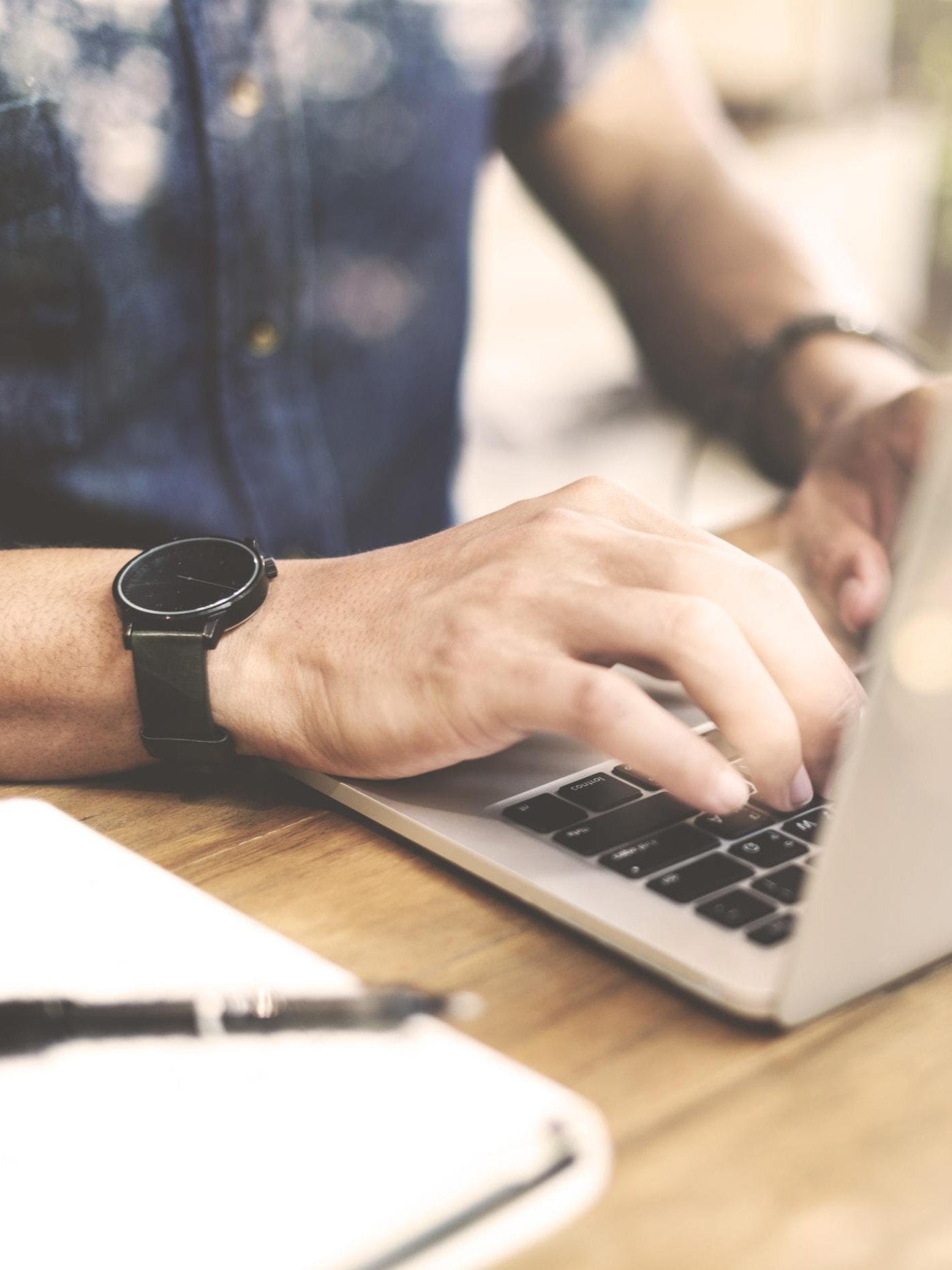 Nahaufnahme von Händen, die auf einer Laptop-Tastatur tippen. Im Vordergrund liegt ein Stift auf einem Notizblock.