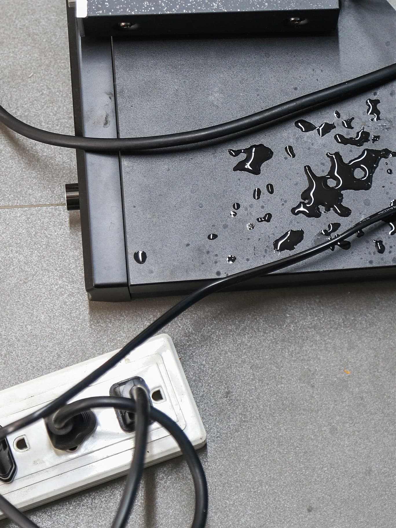 Nasse Stromkabel und eine Steckdosenleiste auf grauem Boden, mit Wassertropfen auf einem schwarzen Gerät, was eine gefährliche Situation darstellt.