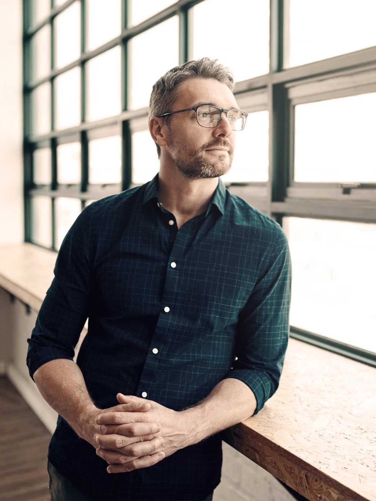 Ein Mann mit Brille und dunkelgrünem Hemd steht in einem modernen Büro und blickt nachdenklich aus einem grossen Fenster mit viel Tageslicht.