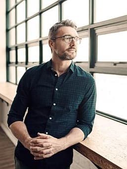 Ein Mann mit Brille und dunkelgrünem Hemd steht in einem modernen Büro und blickt nachdenklich aus einem grossen Fenster mit viel Tageslicht.