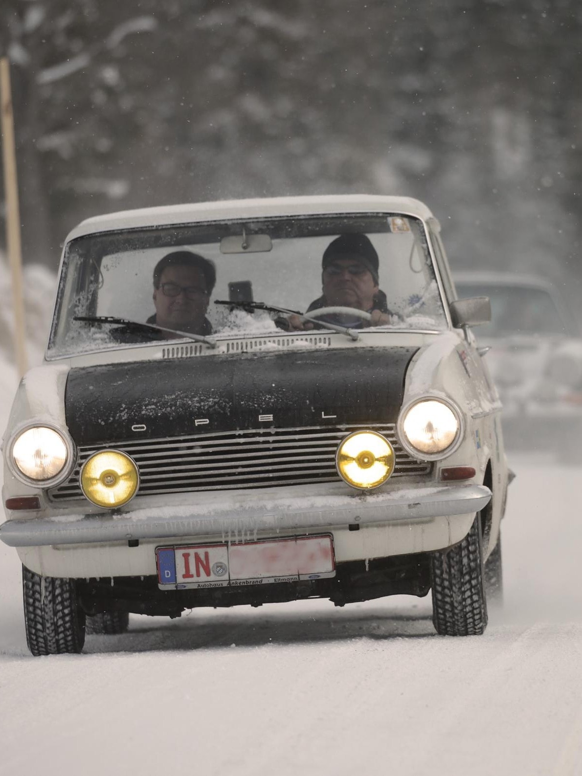 Ein klassisches Ford-Auto fährt auf einer schneebedeckten Straße, gefolgt von einem weiteren Fahrzeug. Die Szene spielt in einem verschneiten Waldgebiet, was auf Winterbedingungen hinweist, und die Scheinwerfer des Autos sind eingeschaltet, und das Nummernschild zeigt teilweise "IN"