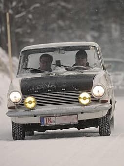 Ein klassisches Ford-Auto fährt auf einer schneebedeckten Straße, gefolgt von einem weiteren Fahrzeug. Die Szene spielt in einem verschneiten Waldgebiet, was auf Winterbedingungen hinweist, und die Scheinwerfer des Autos sind eingeschaltet, und das Nummernschild zeigt teilweise "IN"