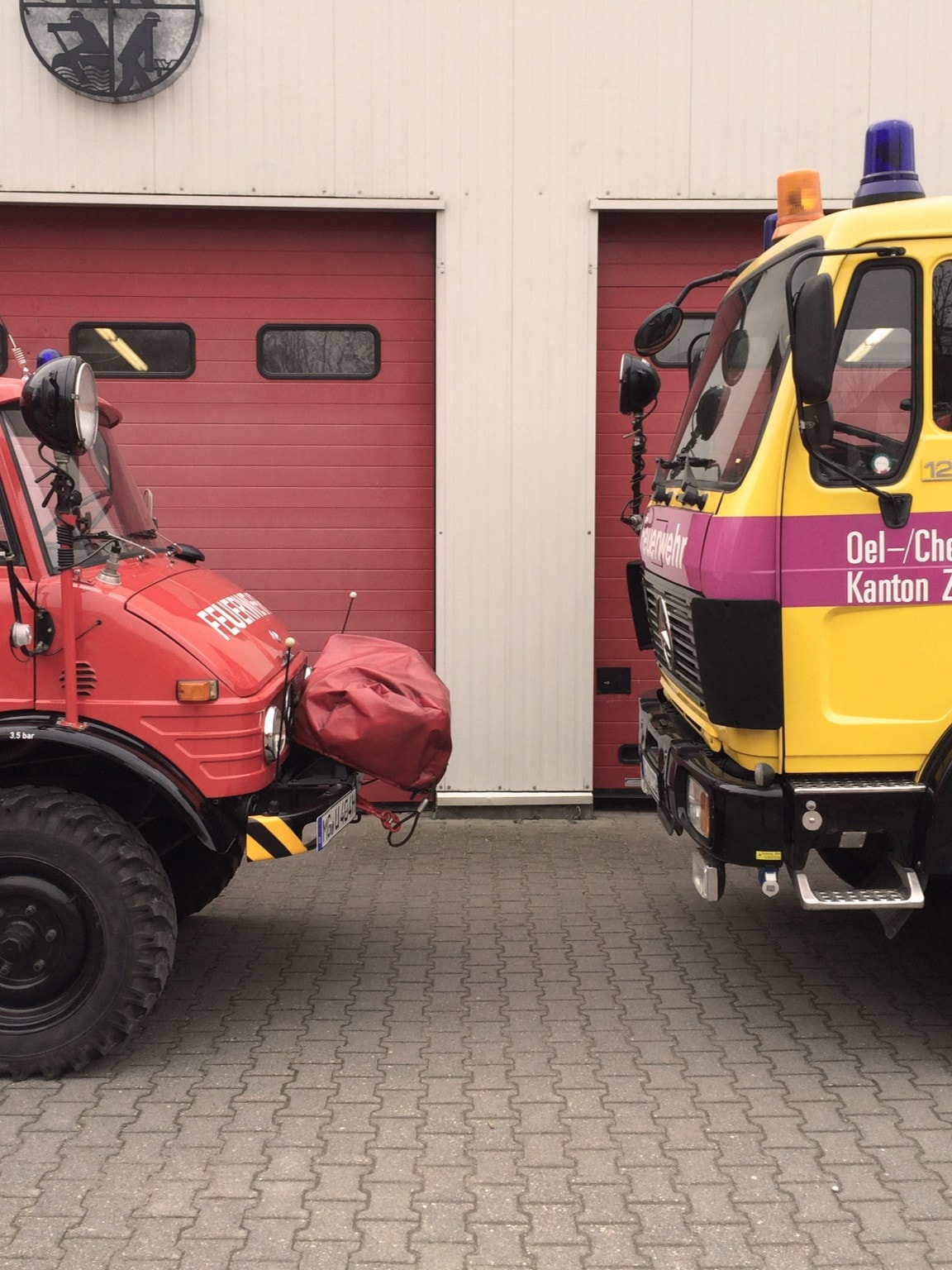 Ein rotes Feuerwehrauto und ein gelbes Feuerwehrauto stehen sich vor einem Gebäude mit roten Garagentoren gegenüber.