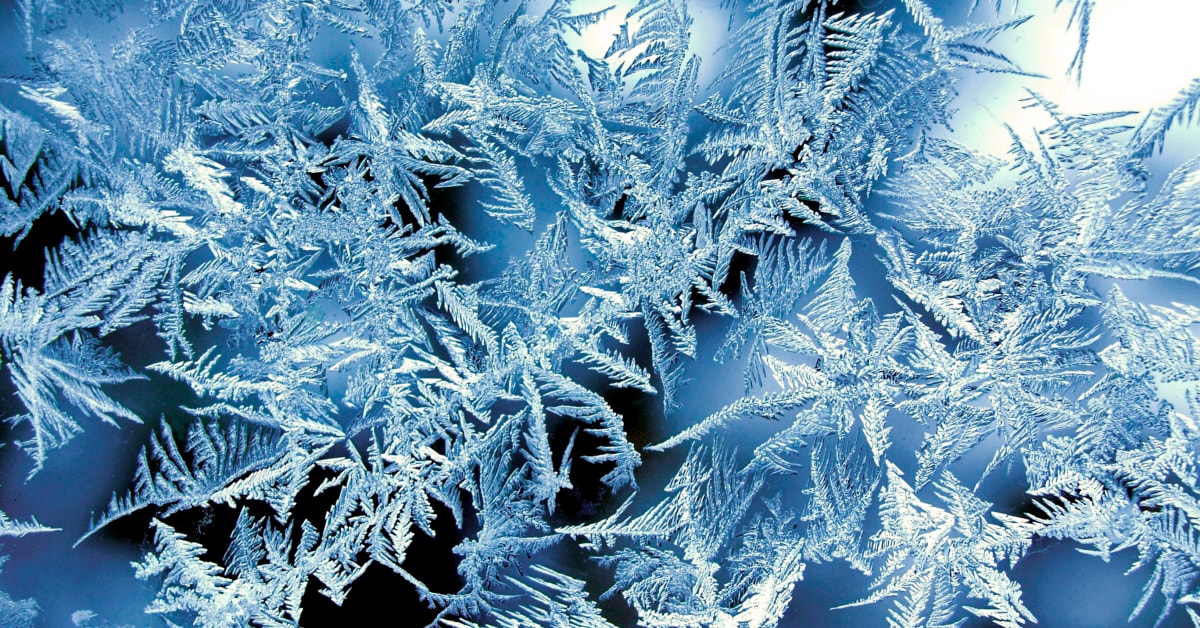 Ein Nahaufnahme von Frostkristallen auf einer Oberfläche. Die Frostkristalle haben komplizierte, federartige Muster und sind von Licht beleuchtet, was eine schöne und detaillierte Darstellung schafft.