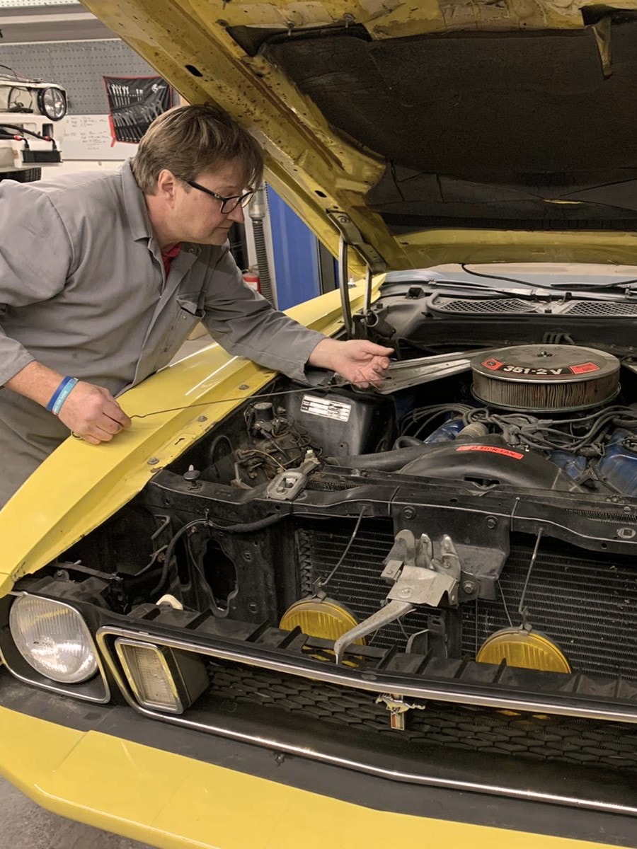 Ein Mechaniker in grauer Arbeitskleidung arbeitet an einem gelben Oldtimer mit geöffneter Motorhaube.