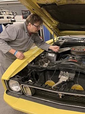 Ein Mechaniker in grauer Arbeitskleidung arbeitet an einem gelben Oldtimer mit geöffneter Motorhaube.