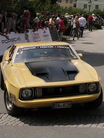 Gelber Ford Mustang fährt an einer Menschenmenge und Werbebannern vorbei während einer Oldtimer-Rallye
