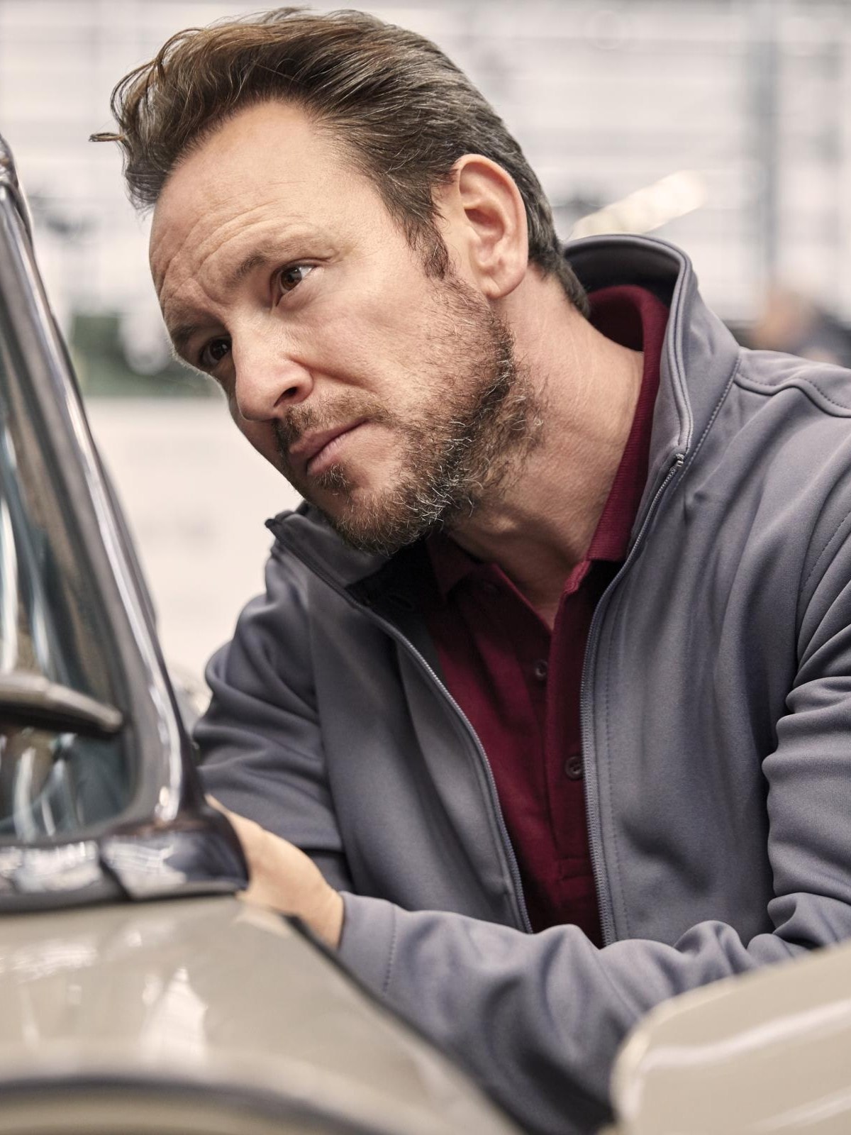 Eine Person mit verschwommenem Gesicht lehnt sich an das Fenster eines klassischen Autos. Die Person trägt eine graue Jacke über einem roten Hemd. Die Szene scheint sich in einer Ausstellung oder einem Showroom abzuspielen.