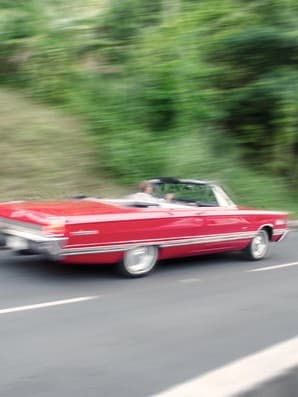 Ein rotes Cabrio fährt auf einer Straße mit verschwommenem Grün im Hintergrund, was auf Bewegung hinweist.