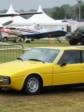 Ein gelber Oldtimer-Sportwagen Simca Bagheera mit schlankem Design fährt auf Gras in der Nähe eines kleinen Flugzeugs und einiger Zelte. Die Szene vermittelt eine nostalgische, klassische Atmosphäre.