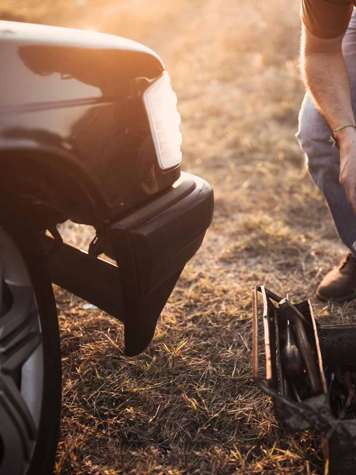 Ein Mann kniet neben einem Auto mit einem offenen Werkzeugkasten und hält Werkzeuge in den Händen.