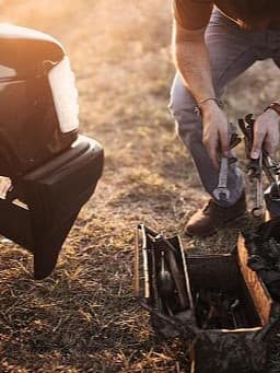 Ein Mann kniet neben einem Auto mit einem offenen Werkzeugkasten und hält Werkzeuge in den Händen.