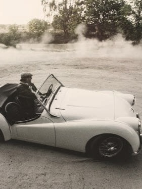 Ein weißes Cabrio driftet auf einer Schotterfläche und wirbelt Staub auf. Im Hintergrund sind Bäume und Grünflächen zu sehen.