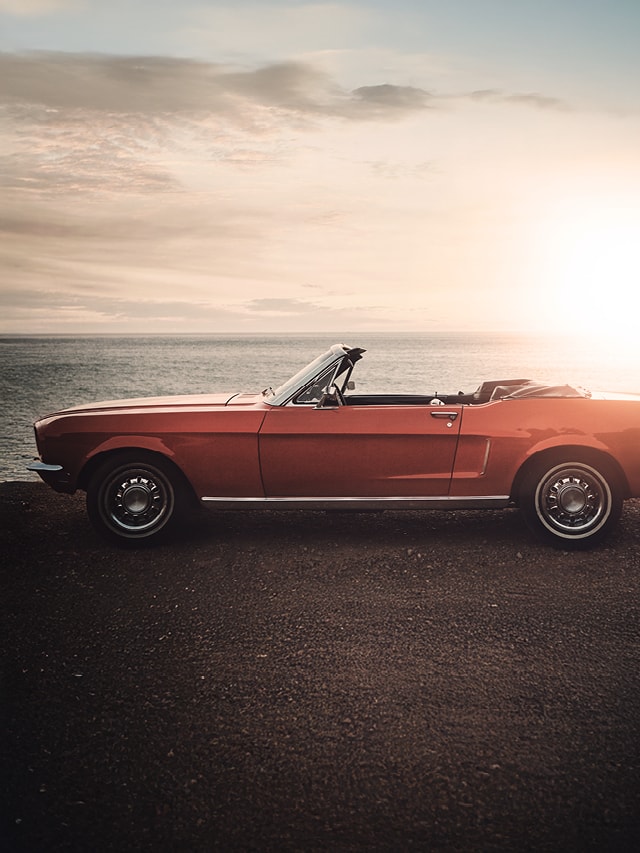 Roter Oldtimer-Cabrio steht seitlich auf einer Küstenstraße mit Blick auf das Meer bei Sonnenuntergang.
