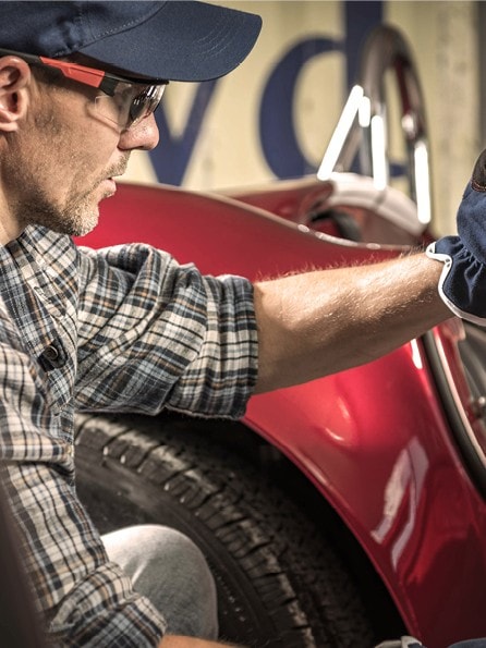 Mann mit Schutzbrile, Cap und kariertem Hemd kniet vor einem rotem Oldtimer und restauriert die Tür des Fahrzeugs